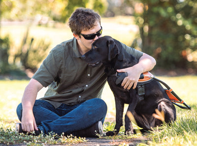 A man with his arm around a Guide Dog while sitting on the grass.
